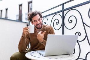 heureux jeune homme souriant, alors qu'il travaille sur son ordinateur portable pour faire toutes ses affaires tôt le matin avec sa tasse de café photo