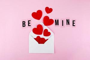 enveloppe blanche, coeurs en papier rouge et texte be mine sur fond rose. saint valentin, concept d'amour photo