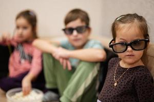 trois enfants assis dans le salon, portent des lunettes 3d en regardant un film ou un dessin animé. photo