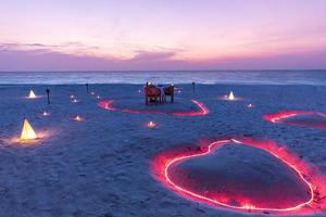 un jeune couple partage un dîner romantique avec des bougies en forme de coeur près de la plage de sable de la mer. belle destination de lune de miel, voyage de luxe, dîner de plage exotique au coucher du soleil avec fond de mer photo