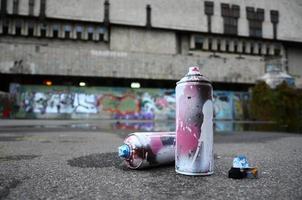 plusieurs bombes aérosols usagées avec de la peinture rose et blanche et des bouchons pour pulvériser de la peinture sous pression se trouvent sur l'asphalte près du mur peint dans des dessins de graffitis colorés photo