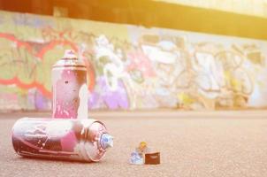 plusieurs bombes aérosols usagées avec de la peinture rose et blanche et des bouchons pour pulvériser de la peinture sous pression se trouvent sur l'asphalte près du mur peint dans des dessins de graffitis colorés photo