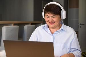 femme âgée heureuse avec casque utilisant un ordinateur portable pour l'éducation, le travail, les achats en ligne, l'écoute de podcasts, etc. photo
