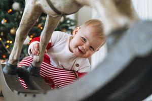 petite fille en costume de père noël rouge regarde par derrière un cheval de bois jouet dans la chambre avec un arbre de noël à la maison photo