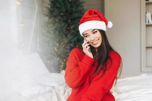 jeune belle femme asiatique aux cheveux longs noirs en pull rouge confortable et bonnet de noel à l'aide d'un smartphone mobile sur le lit dans la chambre avec arbre de noël à la maison photo