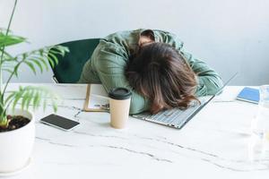 jeune femme brune endormie malheureuse taille plus travaillant sur un ordinateur portable sur une table avec une plante d'intérieur dans le bureau moderne et lumineux photo