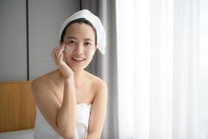 heureuse jeune femme asiatique appliquant des lotions pour le visage tout en portant une serviette et en touchant son visage. maquillage et soins quotidiens photo