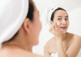 heureuse jeune femme asiatique appliquant des lotions pour le visage tout en portant une serviette et en touchant son visage dans la salle de bain photo