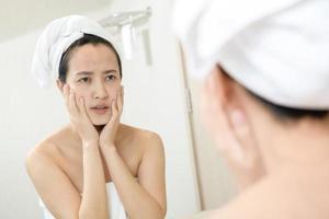 peau à problèmes. concernées les jeunes femmes asiatiques faisant éclater le bouton sur la joue tout en se tenant près du miroir dans la salle de bain. jeunes femmes asiatiques souffrant d'acné photo