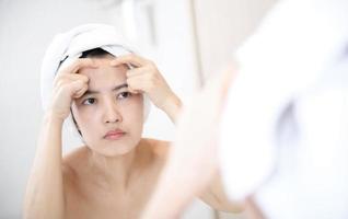 peau à problèmes. concernées les jeunes femmes asiatiques faisant éclater le bouton sur la joue tout en se tenant près du miroir dans la salle de bain. jeunes femmes asiatiques souffrant d'acné photo