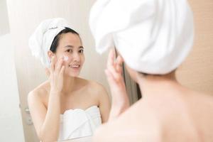 heureuse jeune femme asiatique appliquant des lotions pour le visage tout en portant une serviette et en touchant son visage dans la salle de bain photo