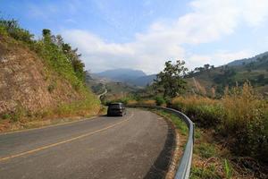 une voiture noire sur une route asphaltée descend et monte sur une colline avec des montagnes et des nuages sur fond de ciel bleu au parc forestier de phu lanka, phayao, thaïlande. beauté du naturel. belle voie, voyage et rue photo