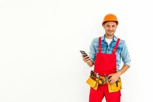 profession, construction et bâtiment - travailleur masculin souriant heureux ou constructeur en casque et dans l'ensemble avec smartphone sur fond blanc photo