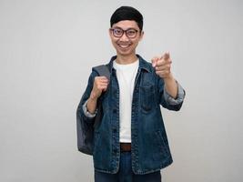 homme asiatique positif chemise jeans porter des lunettes sac de transport sourire et pointer le doigt vers vous isolé photo