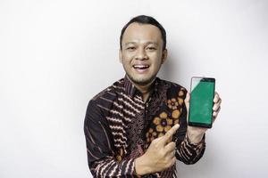 portrait d'un homme asiatique souriant portant une chemise batik et montrant un écran vert sur son téléphone, isolé sur fond blanc photo