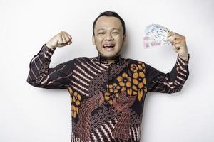 un jeune homme asiatique excité porte une chemise batik et détient de l'argent en espèces en roupie indonésienne gesticulant une pose forte en levant les bras, isolé sur fond blanc photo