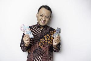 un jeune homme asiatique heureux porte une chemise batik et détient de l'argent en roupie indonésienne isolé sur fond blanc photo