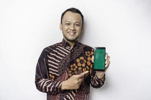 portrait d'un homme asiatique souriant portant une chemise batik et montrant un écran vert sur son téléphone, isolé sur fond blanc photo