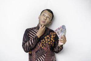 un jeune homme asiatique réfléchi porte une chemise batik et détient de l'argent en espèces en roupie indonésienne isolé sur fond blanc photo