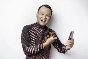 un portrait d'un homme asiatique heureux portant une chemise batik et tenant son téléphone, isolé sur fond blanc photo