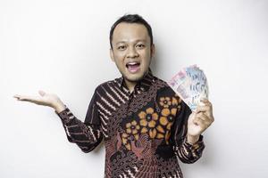 un jeune homme asiatique heureux porte une chemise batik, pointant vers l'espace de copie à côté de lui tout en tenant de l'argent en roupie indonésienne isolé sur fond blanc photo