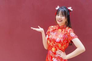 jolie fille asiatique aux cheveux longs qui porte une robe cheongsam rouge sur le thème du nouvel an chinois alors qu'elle montre sa main pour présenter quelque chose sur fond rouge. photo