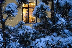fenêtre festive confortable de la maison à l'extérieur avec la lumière chaude des guirlandes lumineuses à l'intérieur - célébrez noël et le nouvel an dans une maison chaleureuse. arbre de noël, bokeh, neige sur les pins et chutes de neige photo