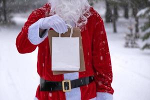 le père noël en plein air en hiver et dans la neige remet des sacs en papier à main avec cadeau artisanal, livraison de nourriture. shopping, recyclage des emballages, fait main, livraison pour noël et nouvel an photo