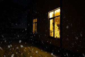 fenêtre festive confortable de la maison à l'extérieur avec la lumière chaude des guirlandes lumineuses à l'intérieur - célébrez noël et le nouvel an dans une maison chaleureuse. arbre de noël, bokeh, neige sur les pins et chutes de neige photo