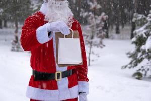 le père noël en plein air en hiver et dans la neige remet des sacs en papier à main avec cadeau artisanal, livraison de nourriture. shopping, recyclage des emballages, fait main, livraison pour noël et nouvel an photo