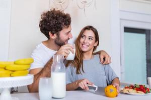 jeune couple souriant cuisinant des aliments dans la cuisine ensemble dans la cuisine, passant un bon moment ensemble. homme et femme riant et buvant du lait le matin avec petit déjeuner photo