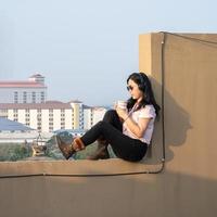 une femme asiatique à lunettes écoute de la musique via un casque, boit du café sur le ciment de la barricade sur le toit pas dangereux car le site opposé est également le sol sur le toit photo