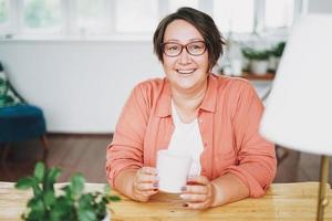 adulte charmante femme brune dans des verres plus la taille du corps positif avec une tasse de thé à la maison photo