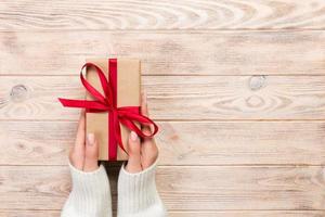 les mains de la femme donnent la saint-valentin enveloppé ou un autre cadeau de vacances fait à la main dans du papier avec un ruban rouge. boîte cadeau, décoration de cadeau sur table en bois blanc, vue de dessus avec espace de copie photo