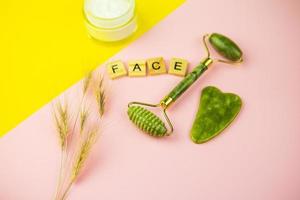 outils de massage facial vert gua sha. rouleau en jade quartz vert sur fond rose-jaune. pot de crème, visage d'inscription en lettres de bois. gros plan, vue de dessus. photo