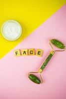 outils de massage facial vert gua sha. rouleau en jade quartz vert sur fond rose-jaune. pot de crème, visage d'inscription en lettres de bois. photo