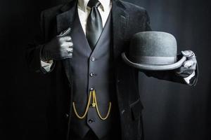 homme en costume sombre et gants en cuir tenant un chapeau melon sur fond noir. concept de gentleman britannique classique et excentrique photo