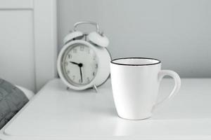 routine du matin à la maison. tasse blanche et réveil sur table de chevet photo