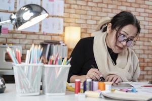 une créatrice de mode asiatique d'âge moyen travaille en studio en dessinant des croquis avec une tablette numérique et un tissu coloré pour une collection de vêtements, une boutique professionnelle tailleur PME entrepreneur. photo