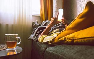 personne allongée sur l'entraîneur à l'aide d'un smartphone avec écran d'espace de copie vierge savourez un thé avec les pieds sur le radiateur dans une maison froide en hiver. système de chauffage central et concept d'économie d'énergie photo