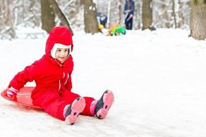 jolie petite fille souriante dans sa combinaison de ski glissant sur une petite colline couverte de neige avec son traîneau photo