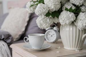 une tasse de café, un réveil et un vase de fleurs blanches sur la table de chevet. l'heure du thé, l'heure du café photo