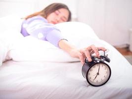 une fille endormie au lit, les yeux fermés, tend la main au réveil à la maison. bonjour un jour paresseux photo