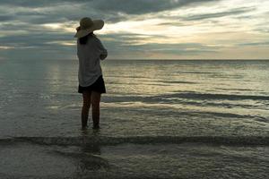 femme portant un chapeau marche seule sur une plage de sable vide au coucher du soleil. une jeune femme solitaire et déprimée se tient sur le sable de la plage en vacances. photo