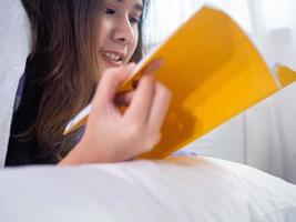 jeune femme est allongée sur le lit en lisant un livre dans la chambre. lire des livres, passer du temps libre pour se détendre pendant les vacances photo