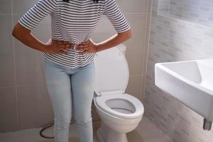 femme se tenait dans la salle de bain dans les toilettes. en cas de diarrhée sévère, utilisez les deux mains pour tenir le ventre, douleur, torsion, torture photo