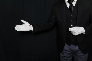 portrait de majordome ou serviteur en costume sombre et gants blancs avec geste de bienvenue sur fond noir. industrie du service. accueil et courtoisie professionnels. photo