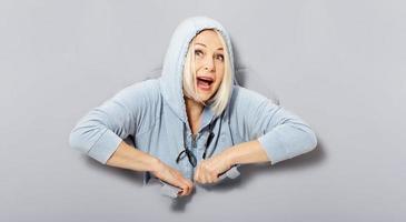 femme joyeuse charismatique avec une coiffure blonde, à l'air ludique, porte un sweat à capuche à la mode, se tient dans un trou de papier déchiré, remarque drôlement agréable. fond gris. photo