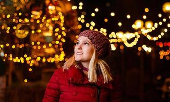 photo en plein air de jeune belle fille souriante heureuse posant dans la rue. foire de noël festive sur fond. modèle portant un manteau d'hiver élégant, un bonnet tricoté, une écharpe.