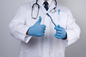 le médecin thérapeute est vêtu d'un uniforme de robe blanche et des gants stériles bleus se tiennent debout et tiennent une brosse à dents photo